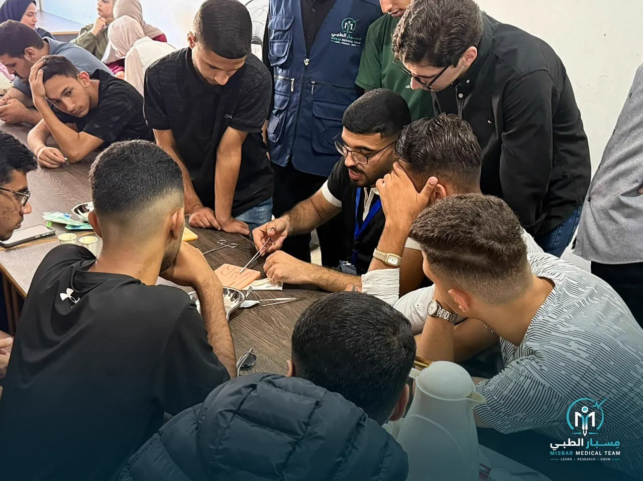 A trainer guides a student’s hands on a suture pad while other students lean in to watch