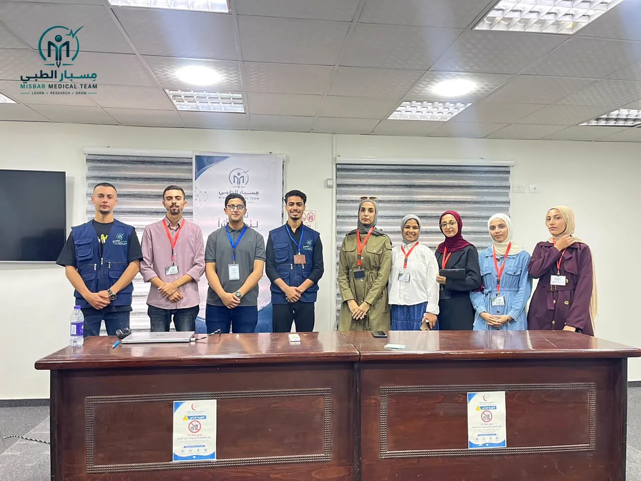 Nine members of the Misbar team standing behind a desk in a lecture room, wearing team vests and badges
