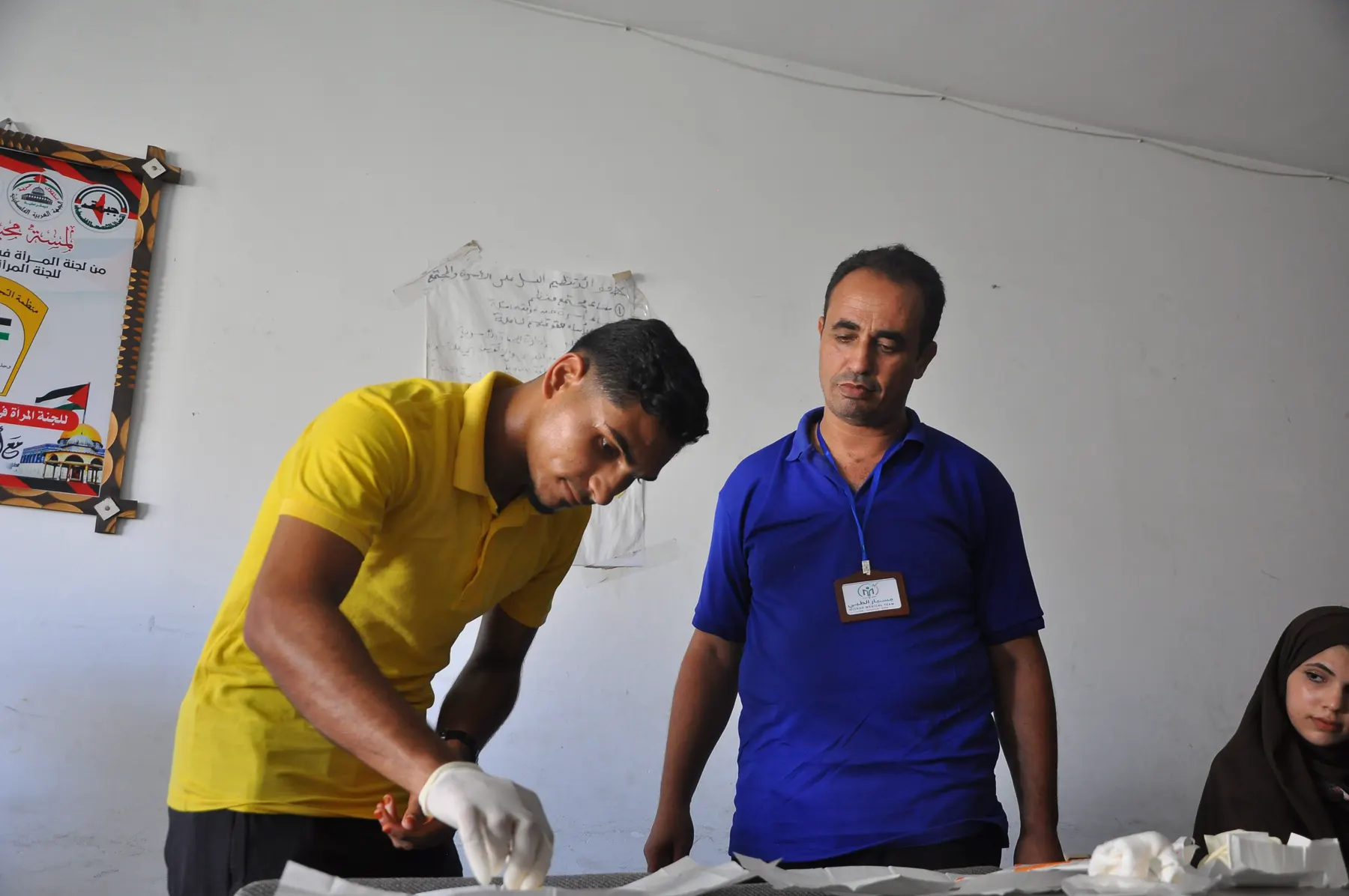 A gloved trainer demonstrates a nursing skill to a student while a supervisor watches